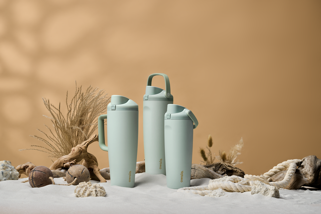 Three teal FreeSip Tumbler bottles in the sand, with shells, rope, and wheat scenic elements