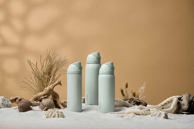Three teal FreeSip bottles in the sand with shells, rope, wheat, and a brown backdrop
